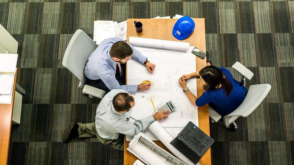 Three people discuss blueprints and construction documents at a table with a blue safety helmet, calculator, and laptop, in a modern office environment.